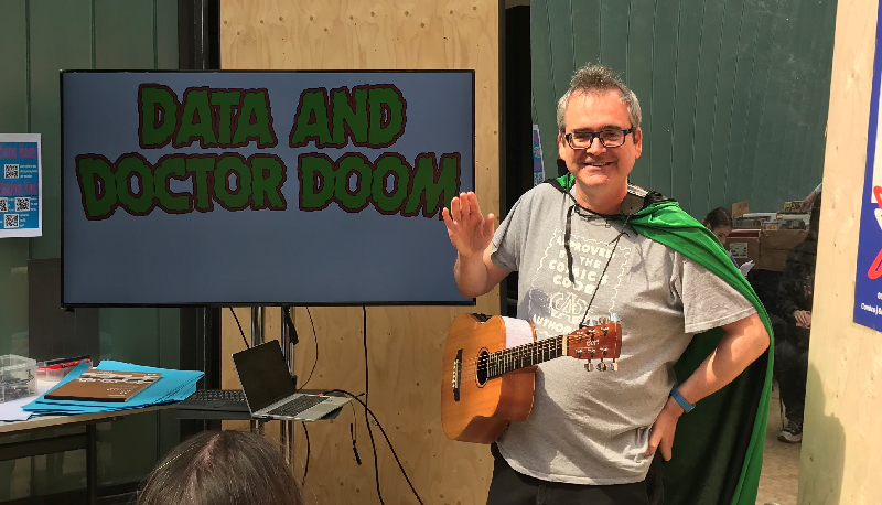 An elegant cape-wearing young man with a guitar around his neck, standing in front of a screen that says Data And Doctor Doom