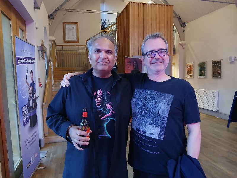 Two impossibly handsome and youthful looking men with their arms around each other in a theatre foyer