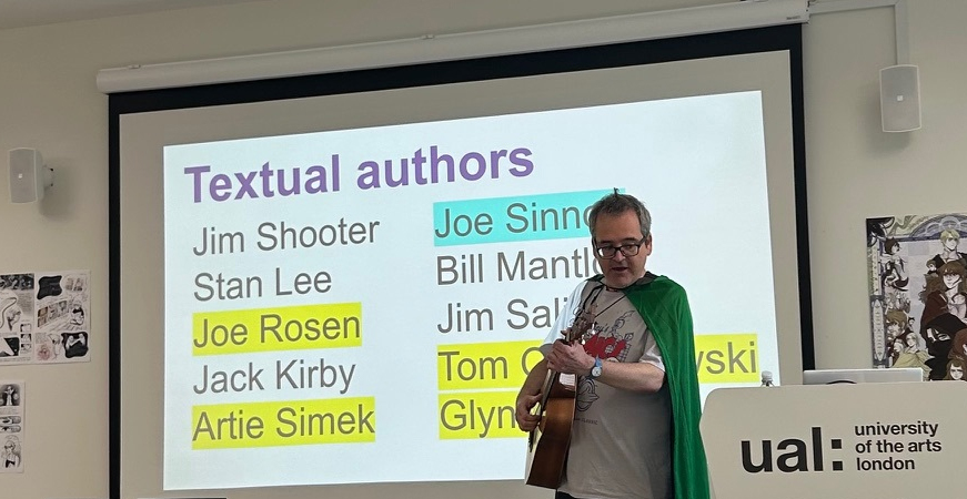 An elegant cape-wearing young man with a guitar around his neck, standing in front of a screen listing the top ten textual authors of Doctor Doom during The Marvel Age